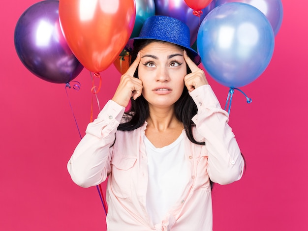 Tevreden jong mooi meisje met een feesthoed die voor ballonnen staat en vingers op ogen zet die op roze muur zijn geïsoleerd