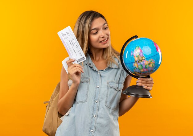 Tevreden jong mooi studentenmeisje die de bol van de achterzakholding en vliegtuigticket dragen die bol bekijken die op oranje muur wordt geïsoleerd