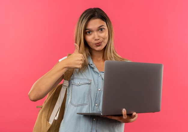 Tevreden jong mooi studentenmeisje die laptop van de achterzakholding dragen en duim tonen die omhoog op roze muur wordt geïsoleerd