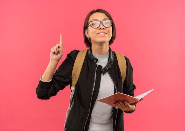 Tevreden jong studentenmeisje die glazen en achterzak met notitieblok dragen die vinger opheffen die op roze muur wordt geïsoleerd