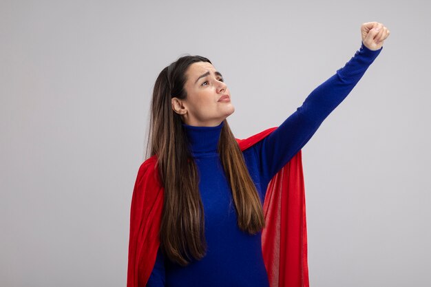 Tevreden jong superheromeisje die kant bekijken die vuist opheffen die op wit wordt geïsoleerd