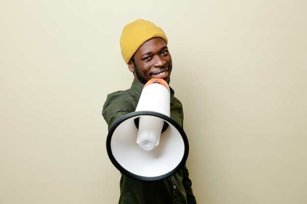 Gratis foto tevreden jonge afro-amerikaanse man in hoed met groen shirt die luidspreker naar camera steekt, geïsoleerd op een witte achtergrond