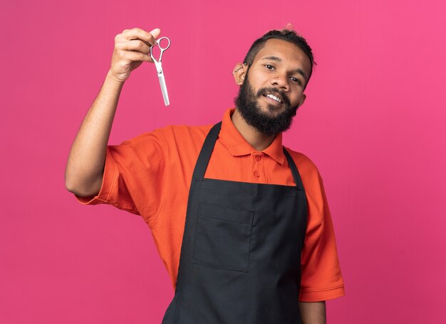 Tevreden jonge Afro-Amerikaanse mannelijke kapper die een uniform draagt met een schaar naar de camera