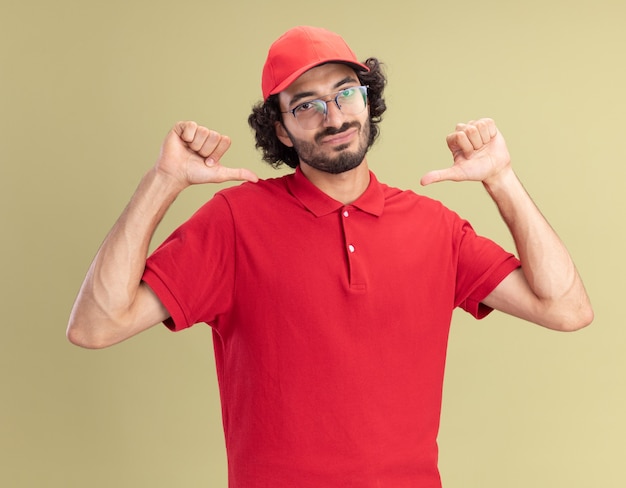 Tevreden jonge bezorger in rood uniform en pet met een bril die naar de voorkant kijkt en naar zichzelf wijst, geïsoleerd op een olijfgroene muur