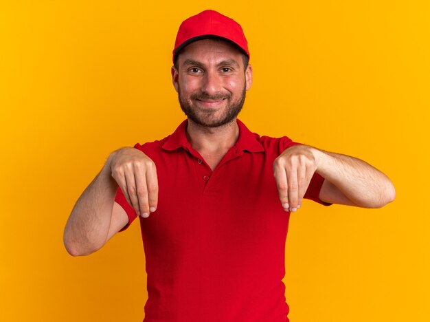 Tevreden jonge blanke bezorger in rood uniform en pet die naar de camera kijkt alsof hij iets op een oranje muur vasthoudt