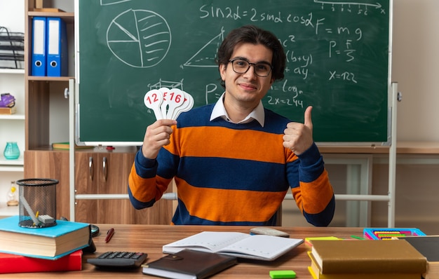 tevreden jonge blanke geometrieleraar met een bril die aan het bureau zit met schoolbenodigdheden in de klas met nummerfans en duim omhoog kijkend naar de voorkant