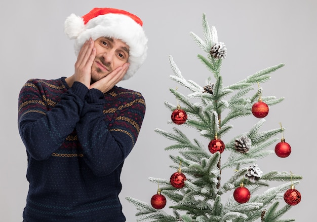 tevreden jonge blanke man met een kerstmuts die in de buurt van de kerstboom staat en zijn handen op zijn gezicht houdt en er geïsoleerd op een witte muur uitziet