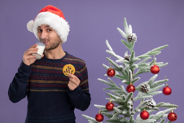 tevreden jonge blanke man met kerstmuts staande in de buurt van versierde kerstboom met glas melk en koekje op zoek naar consumptiemelk geïsoleerd op paarse muur