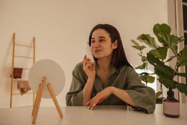 Tevreden jonge blanke vrouw met een frisse huid die een pot crème vasthoudt terwijl ze aan tafel zit