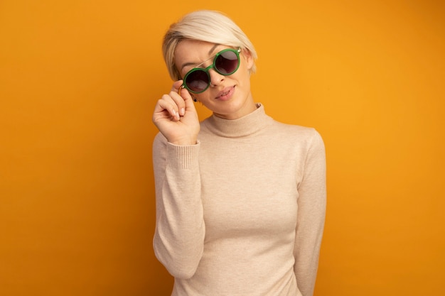 Tevreden jonge blonde vrouw die een zonnebril draagt en grijpt en kijkt naar de voorkant geïsoleerd op een oranje muur met kopieerruimte