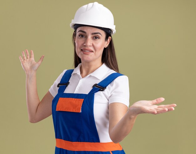 Tevreden jonge bouwersvrouw in eenvormige het uitspreiden die handen op olijfgroene muur worden geïsoleerd