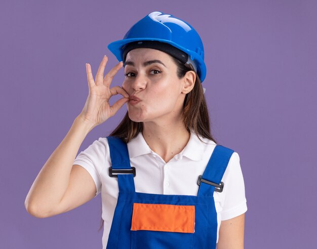 Tevreden jonge bouwersvrouw in uniform die heerlijk gebaar toont dat op purpere muur wordt geïsoleerd