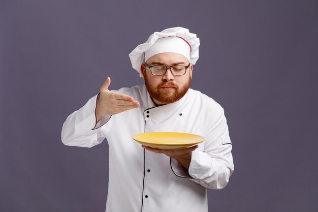 Tevreden jonge chef-kok met een uniforme bril en een pet die een bord vasthoudt en de hand erboven houdt alsof hij een geur van een maaltijd ruikt met gesloten ogen geïsoleerd op een paarse achtergrond