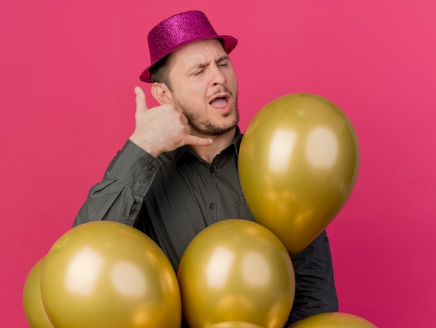 Tevreden jonge feestmens met gesloten ogen die roze hoed dragen die ballons tonen die telefoongesprekgebaar tonen dat op roze wordt geïsoleerd