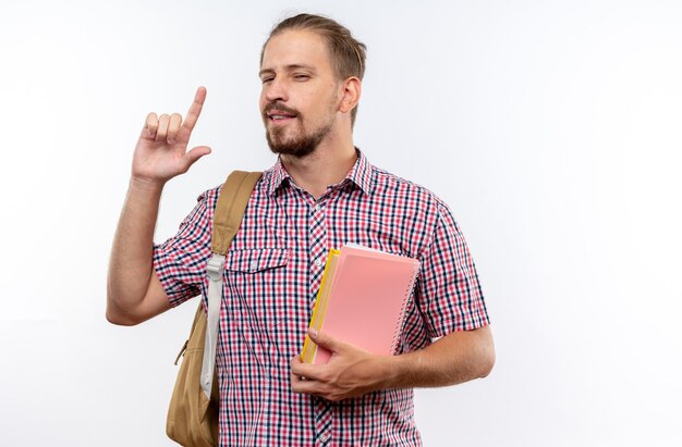 Tevreden jonge kerelstudent die rugzak draagt die boekenpunten op omhoog houdt geïsoleerd op witte muur