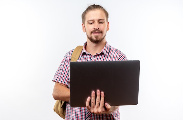 Tevreden jonge kerelstudent die rugzakholding draagt en laptop gebruikt die op witte muur wordt geïsoleerd