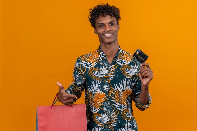 Tevreden jonge knappe donkerhuidige man met krullend haar in bladeren bedrukt overhemd glimlachend met boodschappentassen met creditcard terwijl hij op een oranje achtergrond staat