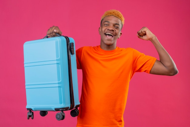 Tevreden jonge knappe jongen die een oranje t-shirt draagt en een reiskoffer vasthoudt glimlachend blij en opgewonden vuist heffen verheugend zijn succes staande over roze muur