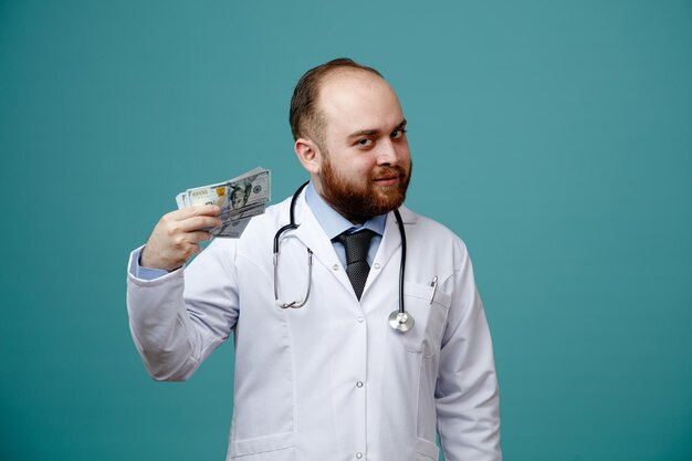 tevreden jonge mannelijke arts die medische jas en stethoscoop om zijn nek draagt en geld laat zien dat naar camera kijkt die op blauwe achtergrond wordt geïsoleerd