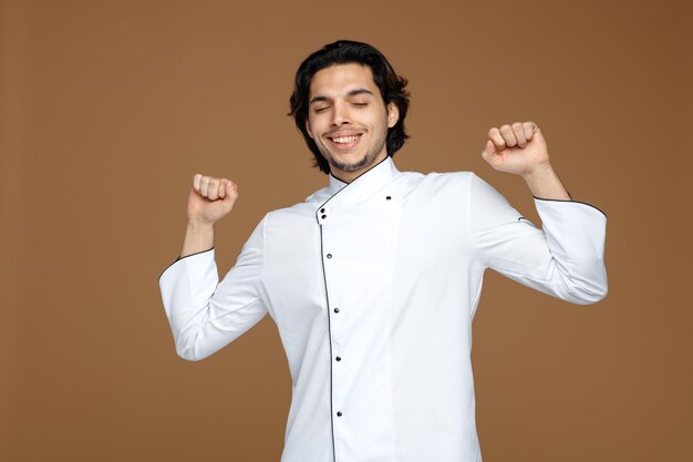 tevreden jonge mannelijke chef-kok die uniform uitrekt en glimlacht met gesloten ogen geïsoleerd op bruine achtergrond