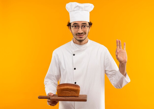 Tevreden jonge mannelijke kok die eenvormige chef-kok en glazen draagt die brood op scherpe raad houdt en okeygebaar toont dat op gele muur wordt geïsoleerd