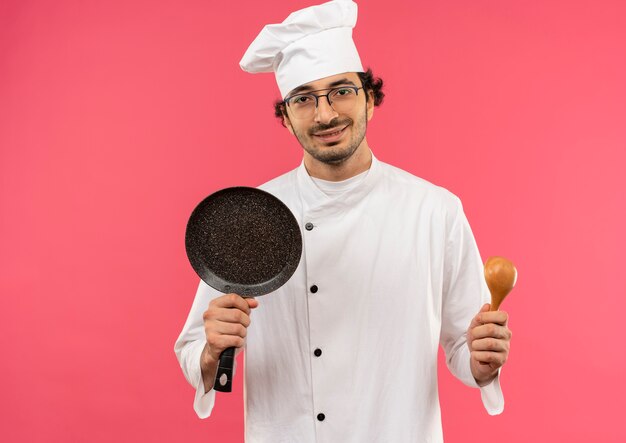 tevreden jonge mannelijke kok die eenvormige chef-kok en glazen draagt die koekenpan en lepel houden