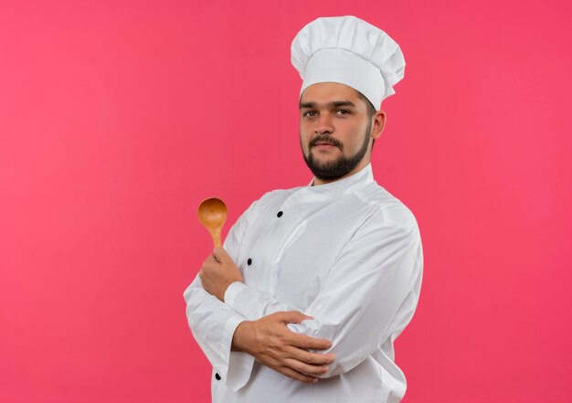 Tevreden jonge mannelijke kok in de lepel van de chef-kok de eenvormige holding en hand op elleboog zetten die op roze ruimte wordt geïsoleerd