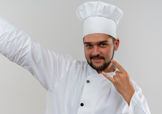 Tevreden jonge mannelijke kok in eenvormige chef-kok die rotsteken doet die uit hand aan kant uitrekt die op witte ruimte wordt geïsoleerd