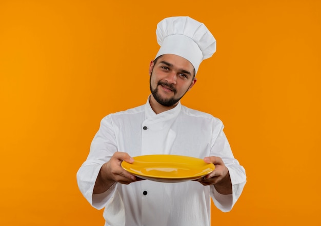 Tevreden jonge mannelijke kok in eenvormige chef-kok die uit lege die plaat uitrekt op oranje ruimte wordt geïsoleerd