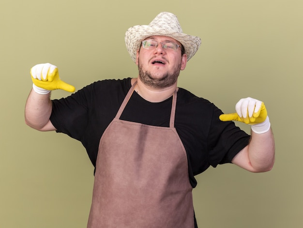 Gratis foto tevreden jonge mannelijke tuinman die een tuinhoed en handschoenen draagt, wijst naar zichzelf geïsoleerd op een olijfgroene muur