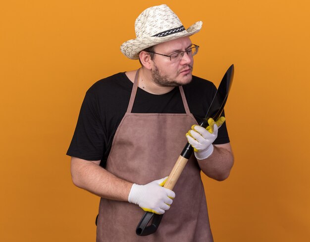 Tevreden jonge mannelijke tuinman die het tuinieren hoed en handschoenen draagt ?? die en schop houdt bekijkt die op oranje muur wordt geïsoleerd