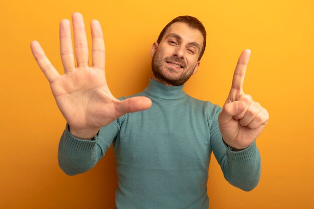 Tevreden jonge mens die voorzijde bekijkt die met zes met handen knipogen toont die op oranje muur worden geïsoleerd