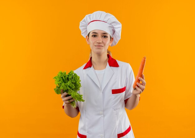 Tevreden jonge mooie kok in de sla en de wortel van de chef-kok de eenvormige die op oranje ruimte wordt geïsoleerd