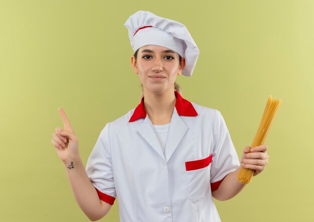 Tevreden jonge mooie kok in de spaghettideegwaren van de chef-kok eenvormige holding en het opheffen van vinger die op groene ruimte wordt geïsoleerd