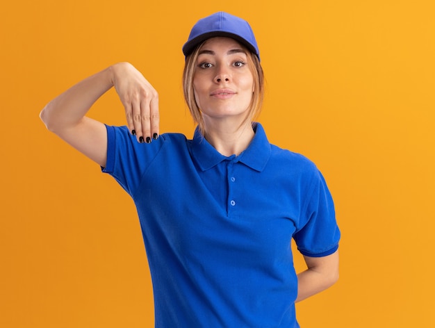 Tevreden jonge mooie leveringsvrouw in uniforme punten naar beneden met hand die op oranje muur wordt geïsoleerd