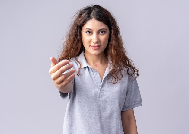 Tevreden jonge mooie vrouw die naar de voorkant kijkt en de hand uitstrekt naar de voorkant geïsoleerd op een witte muur met kopieerruimte