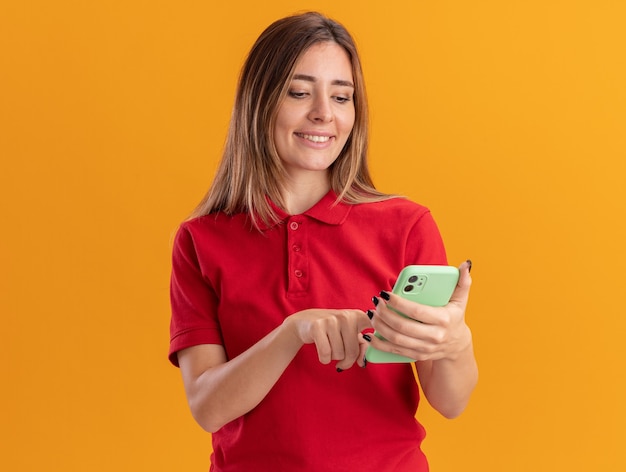 Tevreden jonge mooie vrouw houdt en kijkt naar telefoon geïsoleerd op oranje muur