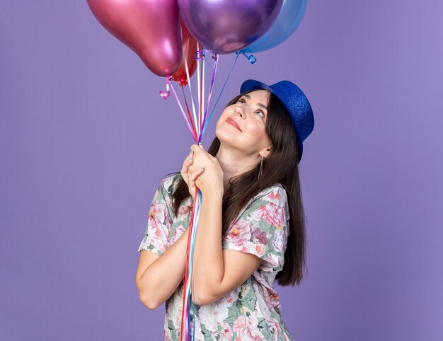 Tevreden jonge mooie vrouw met een feestmuts die ballonnen vasthoudt en bekijkt die op een blauwe muur zijn geïsoleerd