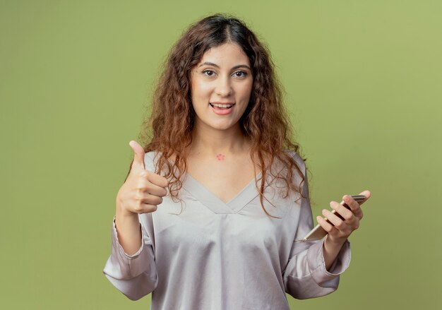Tevreden jonge mooie vrouwelijke kantoormedewerker met telefoon en haar duim omhoog geïsoleerd op olijfgroene muur