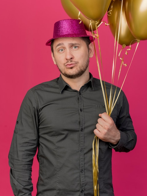 Tevreden jonge partij kerel die roze hoed draagt die ballons houdt die op roze worden geïsoleerd