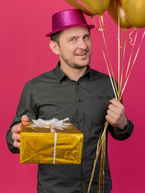 Gratis foto tevreden jonge partij kerel die roze hoed draagt die ballons met giftdoos houdt die op roze wordt geïsoleerd