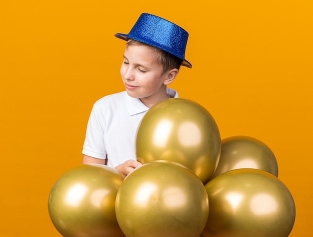 tevreden jonge Slavische jongen met blauwe feestmuts die zich met heliumballons bevindt die op oranje muur met exemplaarruimte worden geïsoleerd