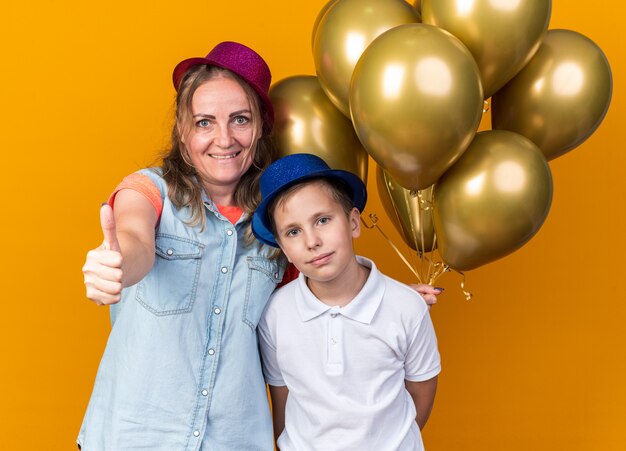 Tevreden jonge Slavische jongen met blauwe feestmuts staan met zijn moeder met paarse feestmuts met helium ballonnen en duim omhoog geïsoleerd op oranje muur met kopie ruimte