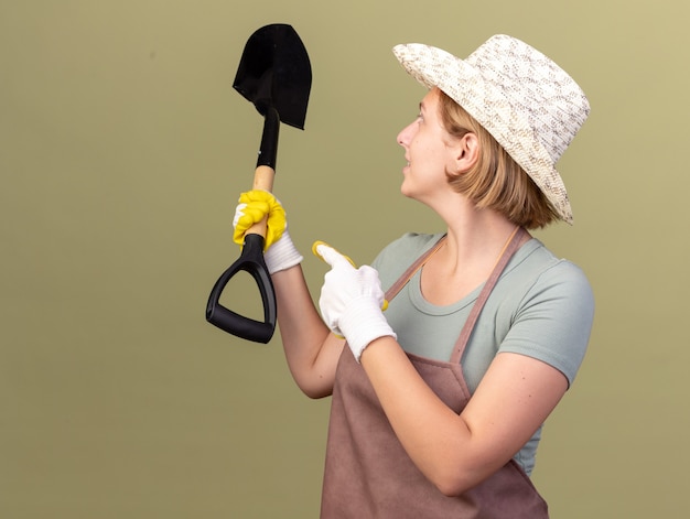 Gratis foto tevreden jonge slavische vrouwelijke tuinman die tuinierende hoed en handschoenen draagt die en op schop op olijfgroen houden richt