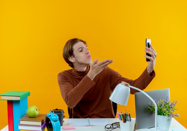 Tevreden jonge studentenjongen die aan bureau met schoolhulpmiddelen zit, neemt een selfie en toont kusgebaar op geel