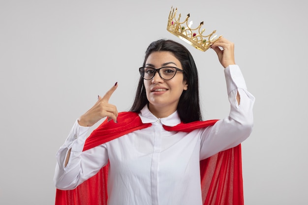 Tevreden jonge supervrouw die een bril draagt die kroon boven het hoofd houdt en naar voorzijde kijkt wijzend op kroon die op witte muur wordt geïsoleerd