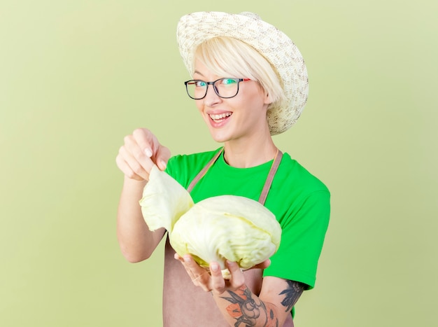 Gratis foto tevreden jonge tuinmanvrouw met kort haar in schort en hoed die kool vrolijk glimlacht