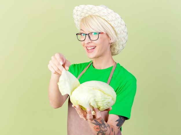 Tevreden jonge tuinmanvrouw met kort haar in schort en hoed die kool vrolijk glimlacht