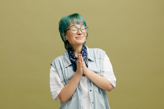 Tevreden jonge vrouw die een glazen bandana op de nek draagt en een namaste-gebaar toont met gesloten ogen geïsoleerd op een olijfgroene achtergrond