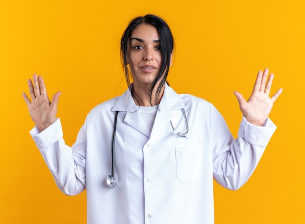 Tevreden jonge vrouwelijke arts die een medisch gewaad draagt met een stethoscoop die handen uitspreidt die op een gele muur zijn geïsoleerd